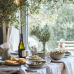 A romantic Mediterranean-inspired dining table with white linens, olive branches, rustic pottery, tzatziki, hummus, pita bread, and Greek wine, set against a pastel green and blue backdrop.